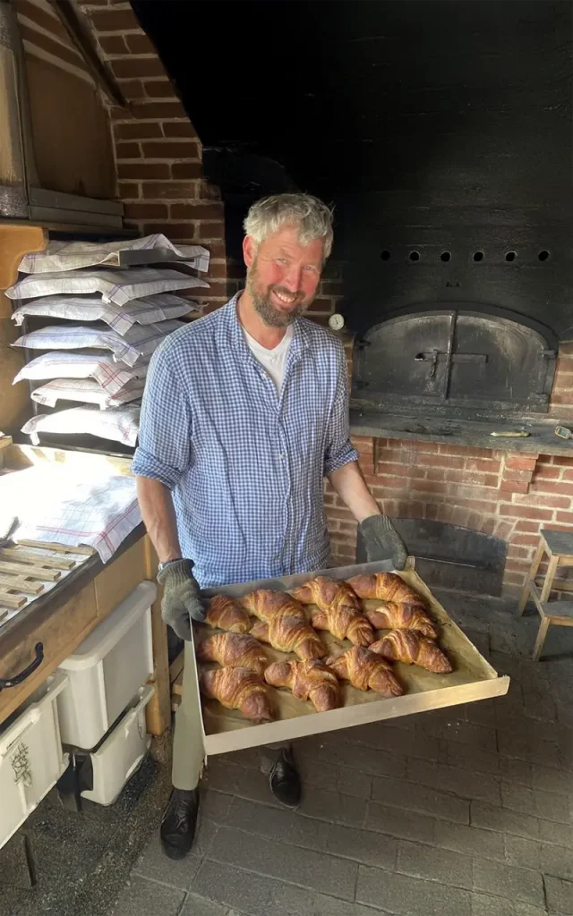 Ein Bäcker hält ein Blech mit frisch gebackenen Croissants vor einem traditionellen Backofen.