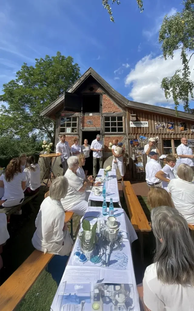 Menschen in weißer Kleidung sitzen an langen Tischen vor einem rustikalen Gebäude im Freien.