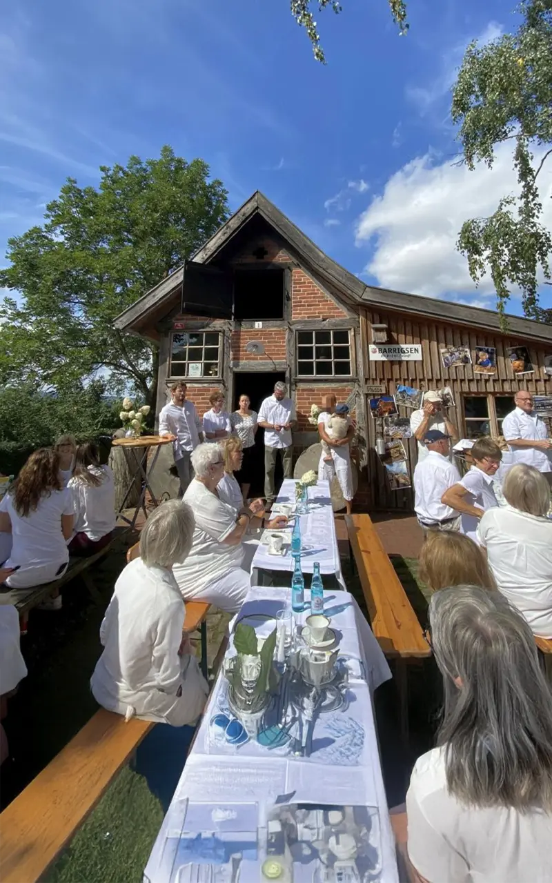 Menschen in weißer Kleidung sitzen an langen Tischen vor einem rustikalen Gebäude im Freien.