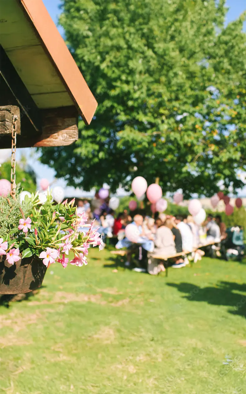 Gartenfeier mit Menschen an Tischen und rosa Luftballons unter Bäumen bei Sonnenschein.